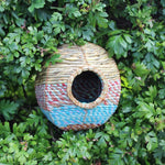 A spherical Anita Artisan Bird Nester woven from natural fibers, featuring blue and red decorative patterns, a round entrance hole, nestled within dense green foliage, providing a cozy and stylish shelter for small garden birds.