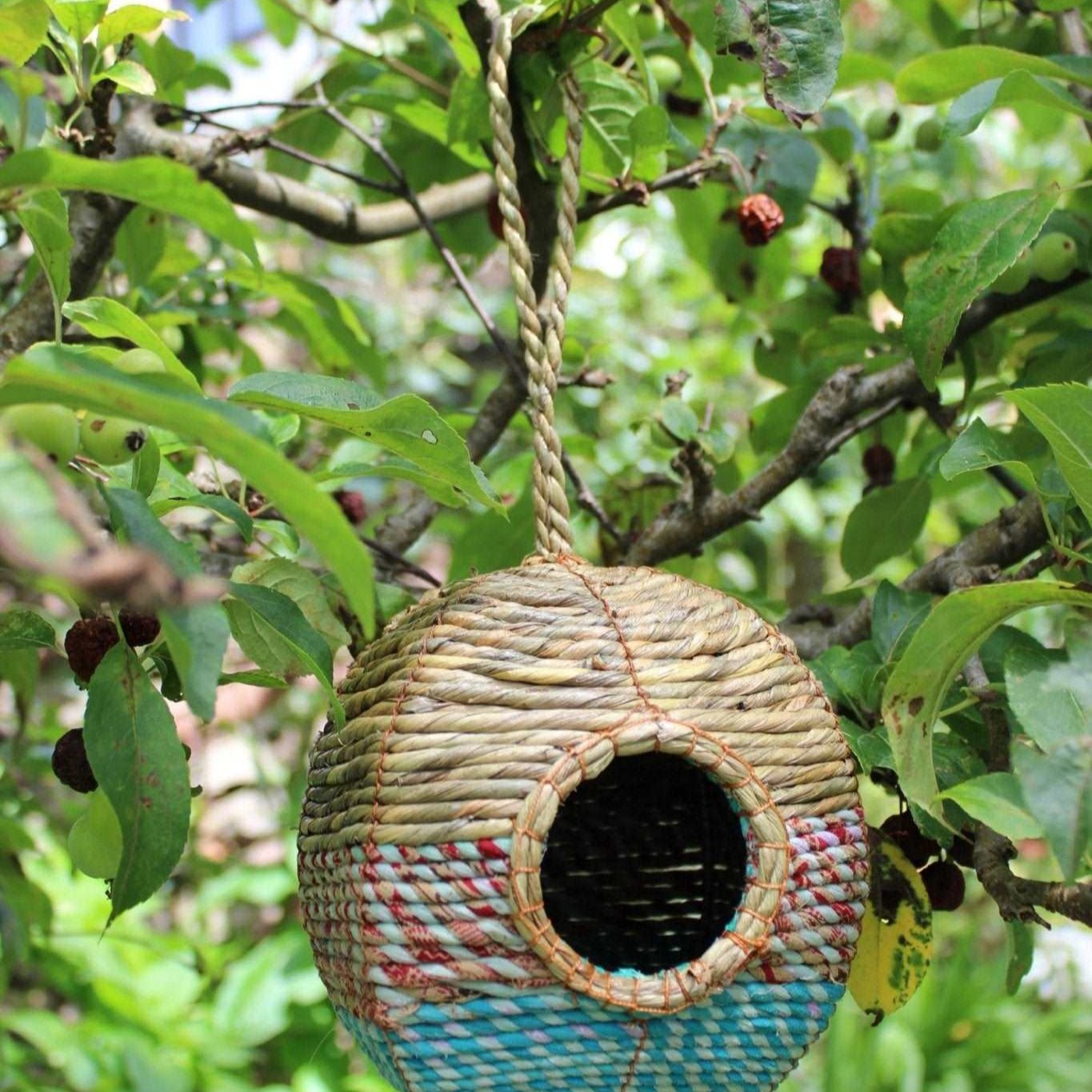 A spherical Anita Artisan Bird Nester woven from natural fibers, featuring colorful blue and red patterns, a round entrance hole, and a sturdy hanging rope, suspended from a tree branch surrounded by lush green foliage, designed as a cozy nesting space for small garden birds.