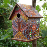 Colourful artisan birdhouse with intricate hand-painted patterns inspired by Bali Island designs, featuring vibrant dots and floral motifs set against a rich brown background, hanging on a tree branch surrounded by lush greenery.