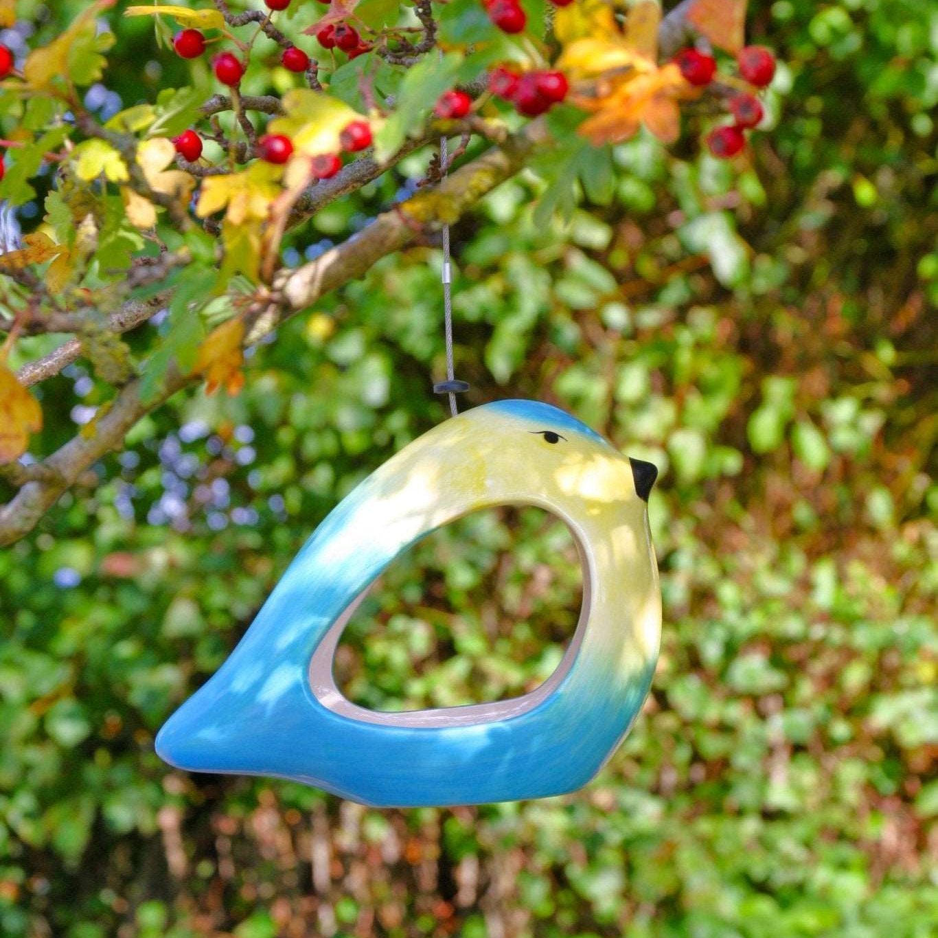 Ceramic Blue Tit Bird Feeder, vibrant blue and yellow bird-shaped feeder, surrounded by green foliage and red berries, hanging from a tree branch, eco-friendly garden accessory