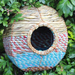 A spherical Anita Artisan Bird Nester, woven from natural materials, featuring colorful blue, red, and beige patterns, a round central entrance hole, securely attached to green foliage, designed as a cozy and decorative shelter for small garden birds.