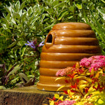 Ceramic Bumblebee Nester, beehive-shaped design, glossy brown ceramic finish, small entrance hole, placed on a garden surface, surrounded by green foliage and colorful flowers, eco-friendly pollinator habitat
