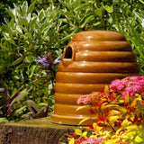 Ceramic Bumblebee Nester, beehive-shaped design, glossy brown ceramic finish, small entrance hole, placed on a garden surface, surrounded by green foliage and colorful flowers, eco-friendly pollinator habitat