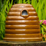 Ceramic Bumblebee Nester, beehive-shaped design, glossy brown ceramic finish, small entrance hole, placed on a garden surface, surrounded by green foliage and colorful flowers, eco-friendly pollinator habitat