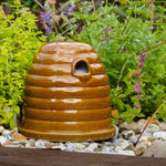 Ceramic Bumblebee Nester, beehive-shaped design, glossy brown ceramic finish, small entrance hole, placed on a garden surface, surrounded by green foliage and colorful flowers, eco-friendly pollinator habitat