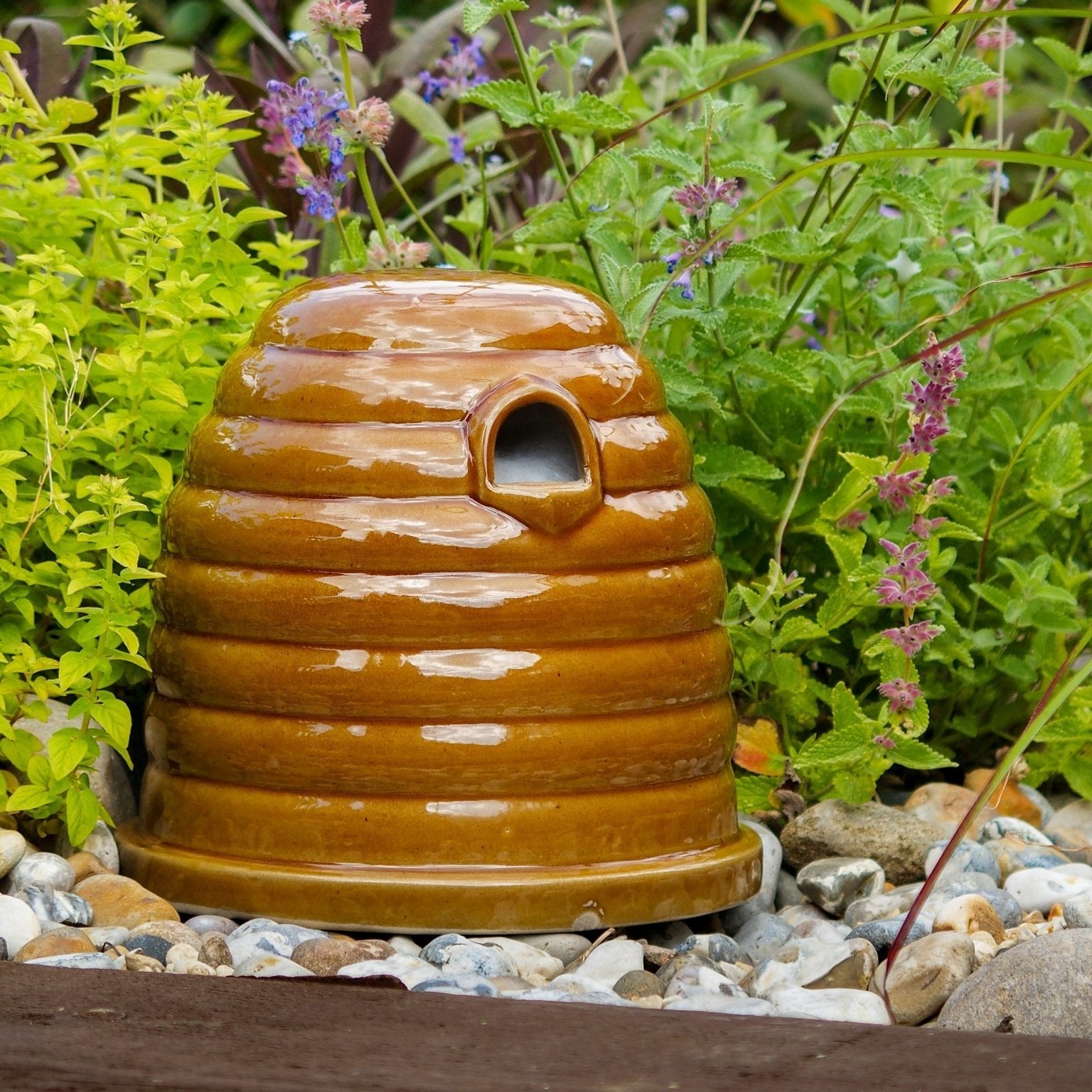 Ceramic Bumblebee Nester, beehive-shaped design, glossy brown ceramic finish, small entrance hole, placed on a garden surface, surrounded by green foliage and colorful flowers, eco-friendly pollinator habitat
