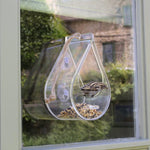 Transparent dewdrop-shaped window bird feeder, made of clear acrylic, attached to a window with suction cups, filled with birdseed, featuring a small bird perched inside, rope accent for decorative touch, modern bird feeder design, surrounded by blurred greenery in the background