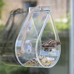 Dewdrop Window Bird Feeder - The Irish Gardener Store Transparent dewdrop-shaped window bird feeder, made of clear acrylic, attached to a window with suction cups, filled with birdseed, featuring a small bird perched inside, rope accent for decorative touch, modern bird feeder design, surrounded by blurred greenery in the background
