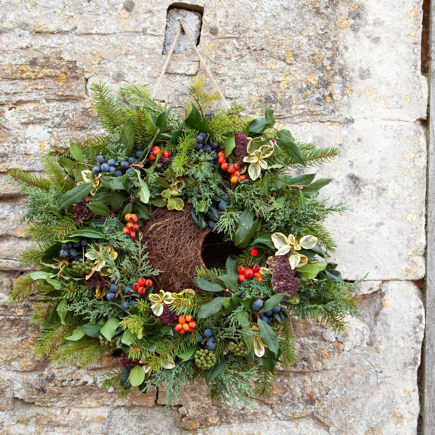 Simon King Wreath Nester - The Irish Gardener Store Simon King Wreath Nester hanging on a stone wall, adorned with natural foliage and berries, eco-friendly bird nester, decorative wildlife habitat, rustic garden feature, sustainable nesting solution for small birds, handcrafted wreath design, outdoor bird-friendly accessory, vibrant greenery and colorful berries