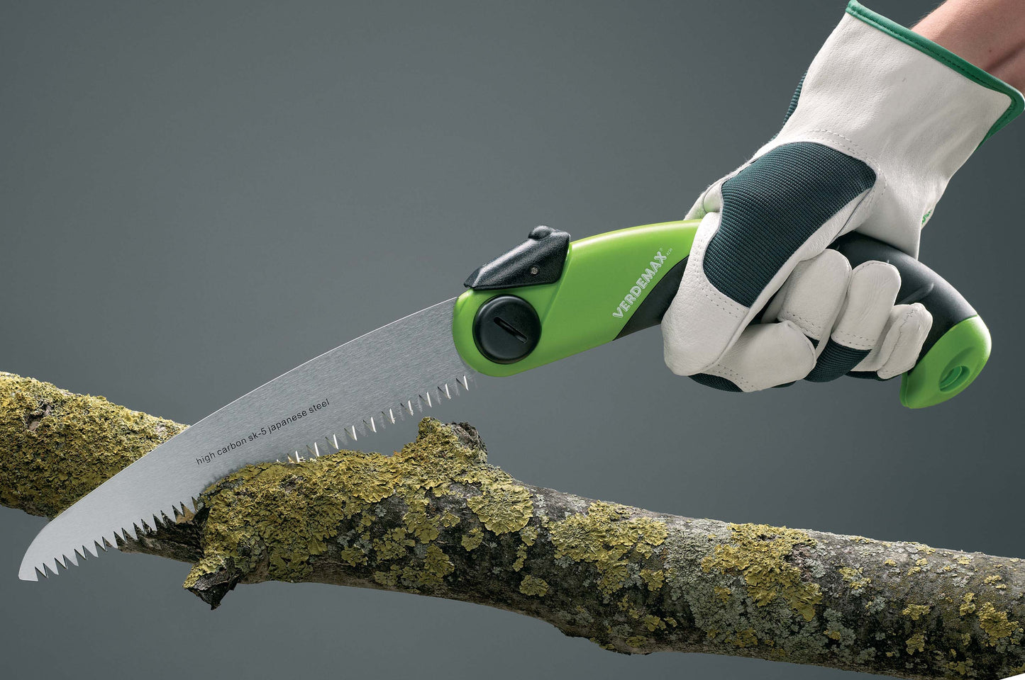 A person using the Verdemax Folding Saw to cut through a branch. The saw features a green and black ergonomic handle, with the blade made of high carbon SK-5 Japanese steel. The cutting teeth are designed for effective, powerful cuts. The folding mechanism ensures easy storage and portability.