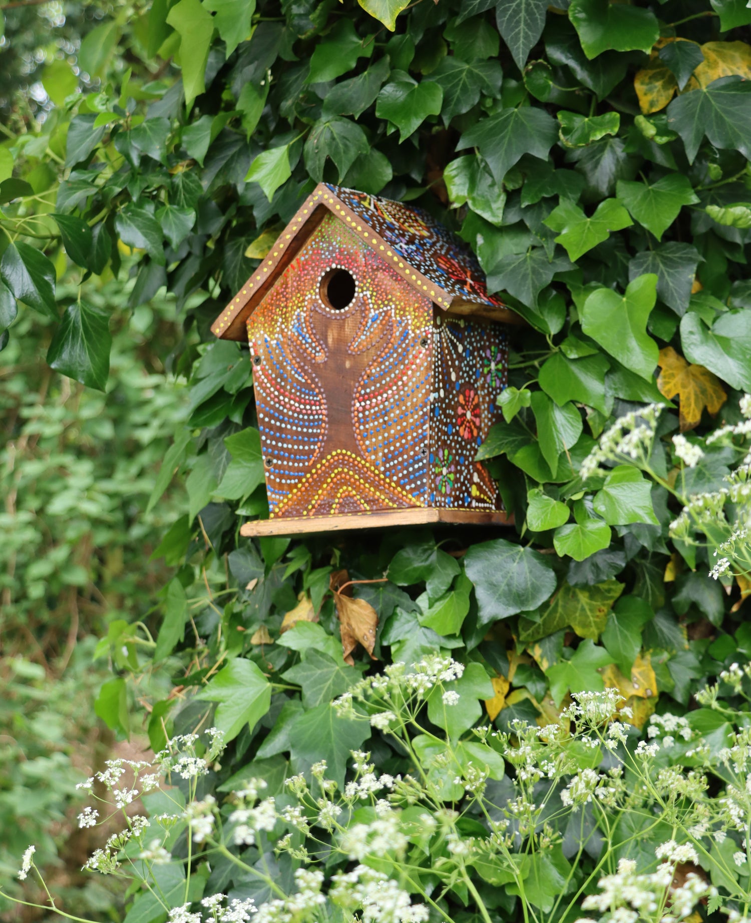 Colourful artisan birdhouse with intricate hand-painted patterns inspired by Bali Island designs, featuring vibrant dots and floral motifs set against a rich brown background, hanging on a tree branch surrounded by lush greenery.