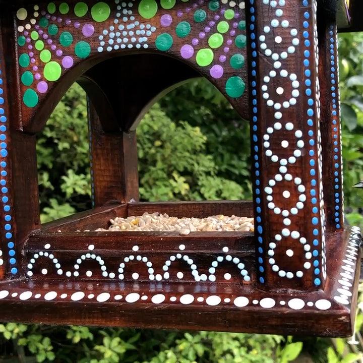 **Alt Text:** A beautifully hand-painted Artisan Hanging Bird Table from the Bali Island Range, featuring intricate dot-painting patterns in white, blue, green, and purple on dark wooden panels. The table is filled with bird seed, and a small bird is perched on the edge, ready to feed. The lush green garden background enhances the natural and artistic design.