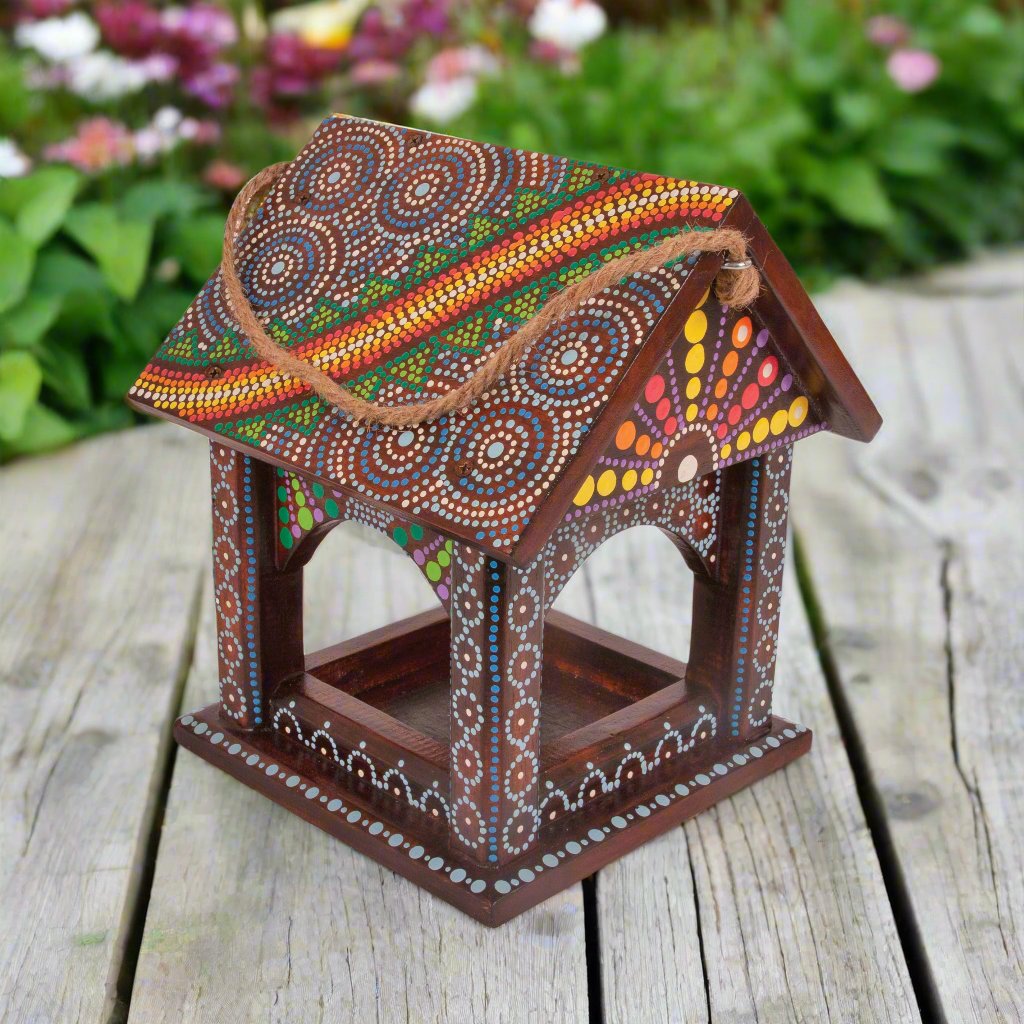 A handcrafted Artisan Hanging Bird Table from the Bali Island Range. The wooden bird table features vibrant hand-painted dot patterns in red, yellow, white, and blue, with intricate details covering the roof and frame. 