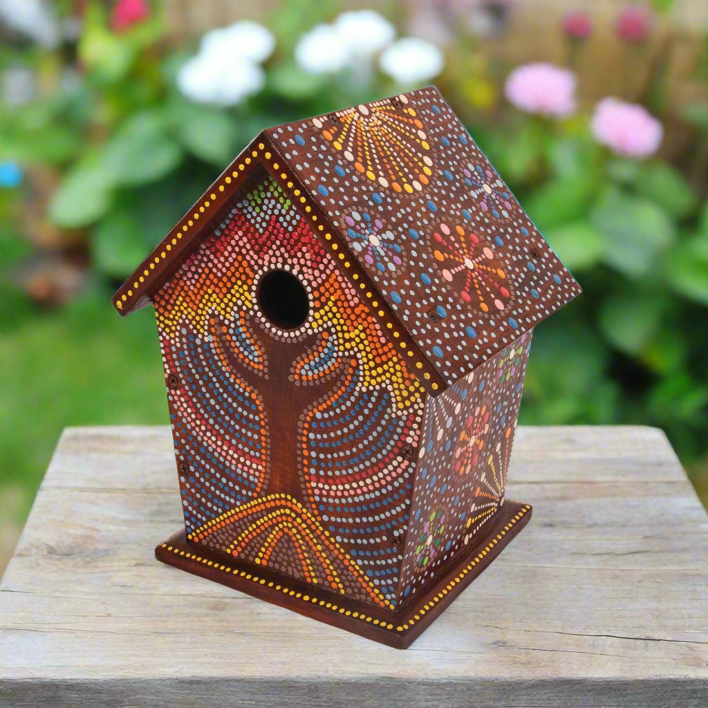 Colourful artisan birdhouse with intricate hand-painted patterns inspired by Bali Island designs, featuring vibrant dots and floral motifs set against a rich brown background, hanging on a tree branch surrounded by lush greenery.