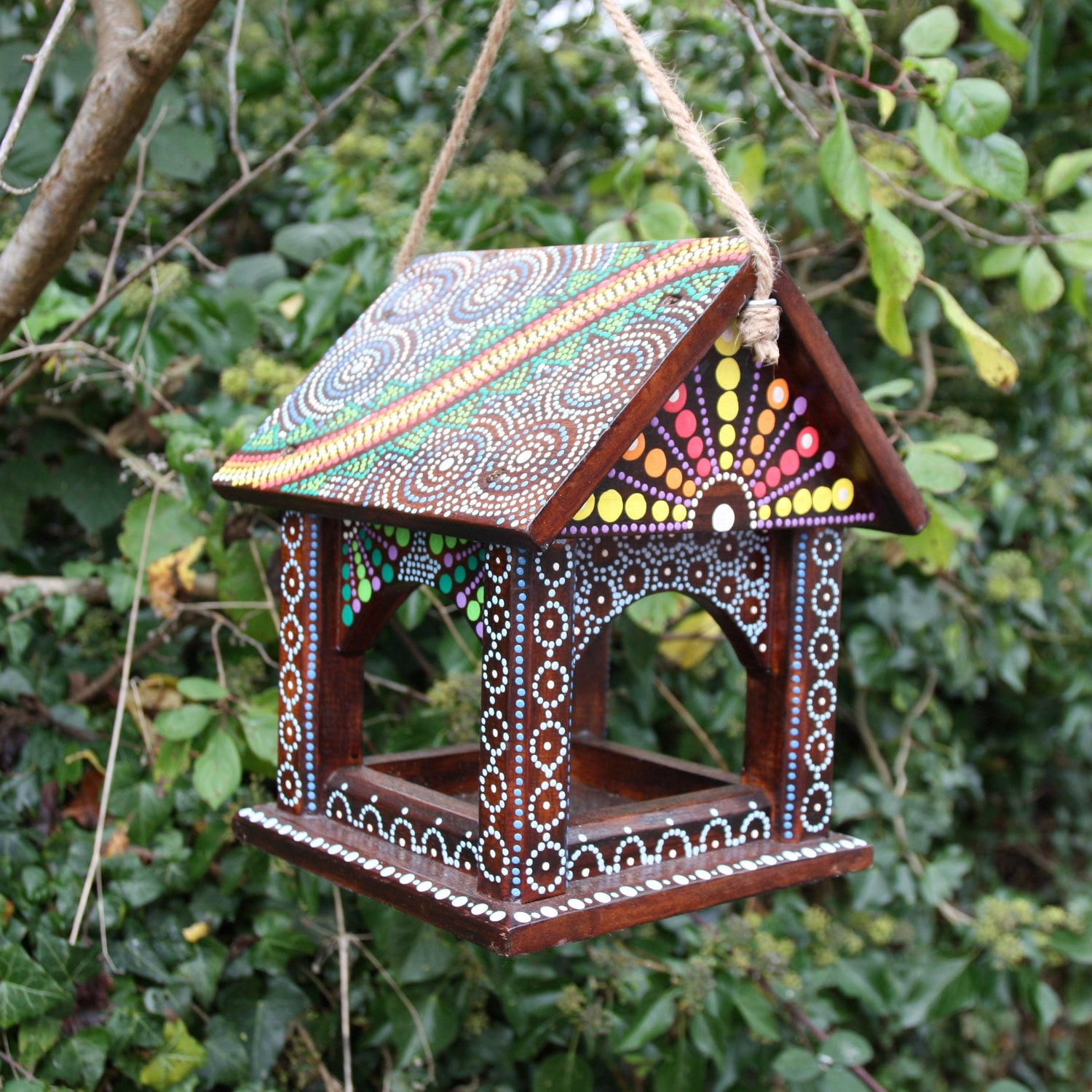 A vibrant Artisan Hanging Bird Table from the Bali Island Range, suspended by a rope in a lush green garden. The table is handcrafted from wood and adorned with intricate dot-painting patterns in white, blue, red, yellow, green, and orange, covering the roof and sides. The colorful design and detailed craftsmanship reflect traditional Balinese artistry.