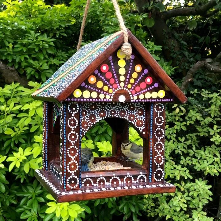 A handcrafted Artisan Hanging Bird Table from the Bali Island Range, suspended by a rope in a lush green garden. The wooden bird table features vibrant hand-painted dot patterns in red, yellow, white, and blue, with intricate details covering the roof and frame. Two small birds are perched inside the table, feeding on seeds, adding a natural charm to the scene.