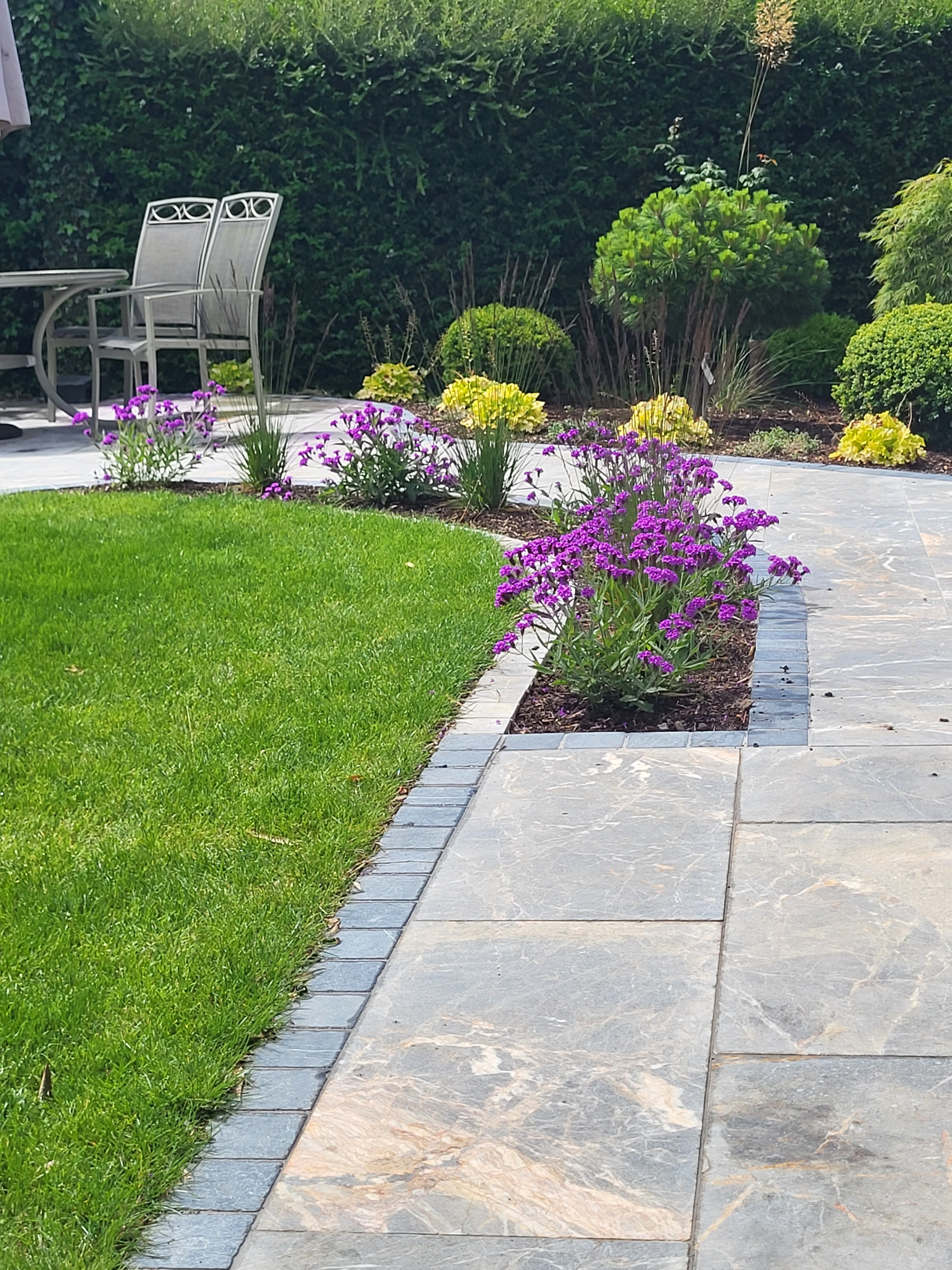 "Beautifully designed modern garden with a curved lawn, paved pathway, and vibrant purple and yellow flowering plants. A perfect outdoor space with structured planting and elegant patio seating.
