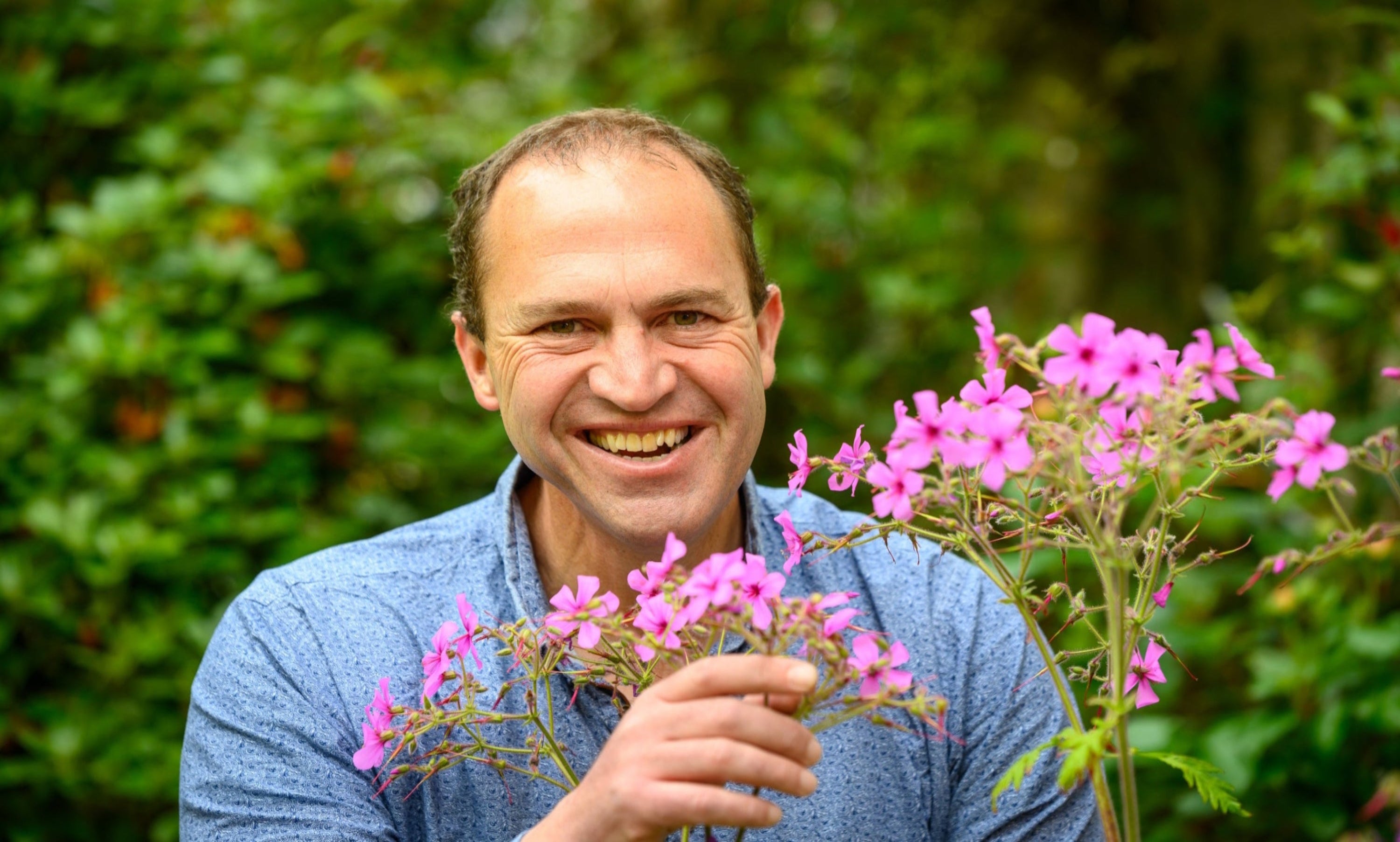 Peter Dowdall, expert garden designer, smiling while holding vibrant pink flowers, set against a lush green garden background.