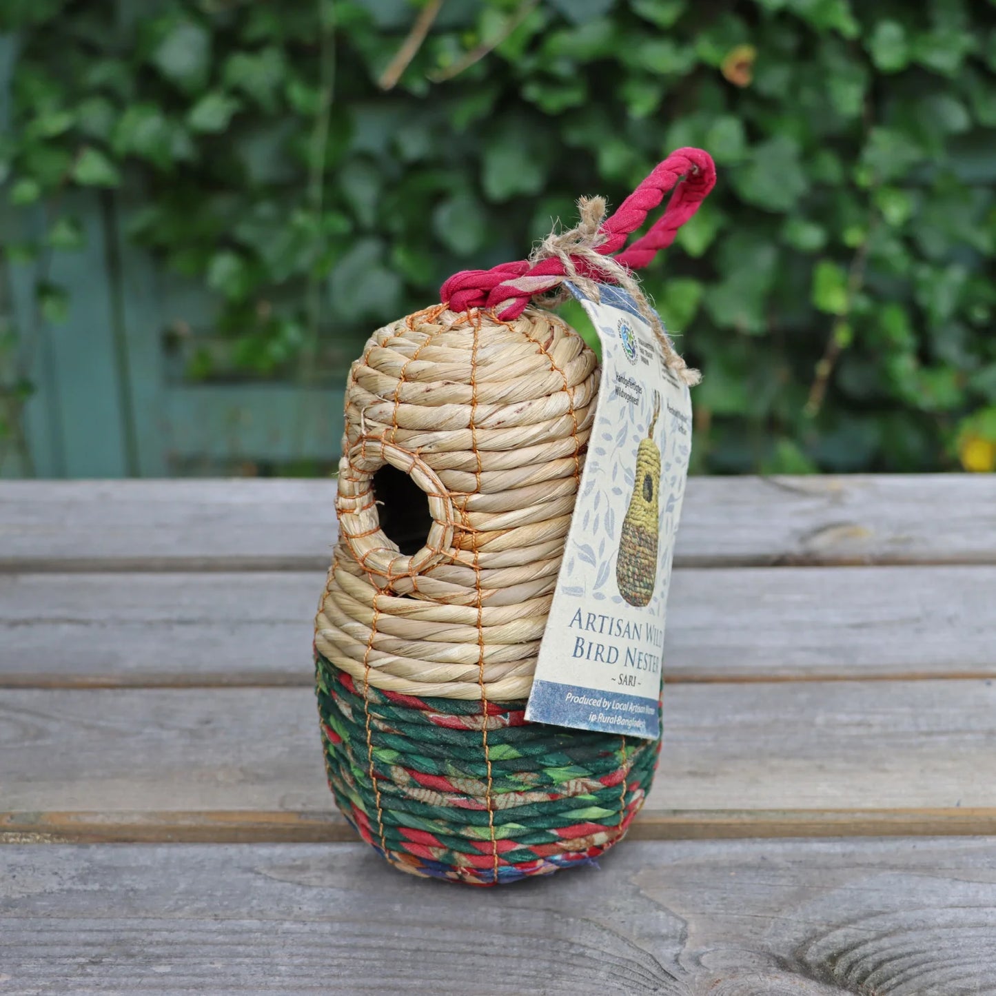 Close-up of the Sari Artisan Bird Nester on a wooden surface, showcasing its natural woven design in beige, green, and red hues. A sturdy red rope is attached for hanging, with an eco-friendly tag highlighting its handmade craftsmanship. Ideal for sustainable garden decor and providing shelter for small garden birds.