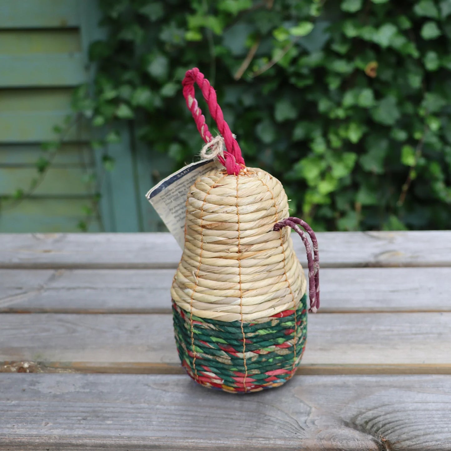 Back view of the Sari Artisan Bird Nester showcasing its natural woven design in beige, green, and red tones. The eco-friendly birdhouse features a sturdy red hanging rope and additional hooks for easy attachment. Perfect for sustainable garden decor and creating a cozy nesting space for small birds