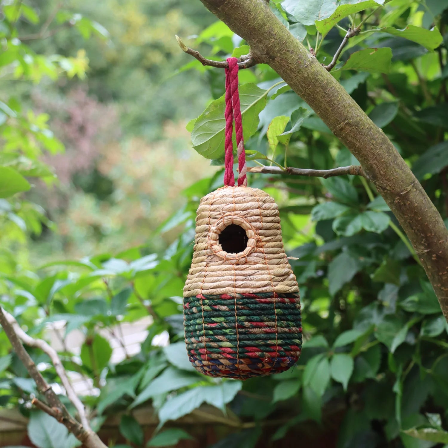 Handcrafted Sari Artisan Bird Nester hanging from a tree branch, featuring its natural woven design with beige, red, and green hues. The birdhouse includes a sturdy red hanging rope and visible attachment hooks, surrounded by lush green foliage. Ideal for eco-friendly garden decor and a safe nesting spot for small birds