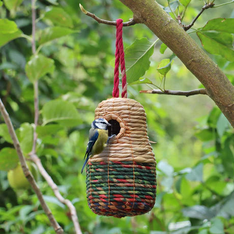 Sari Artisan Bird Nester hanging from a tree branch, handcrafted with natural woven materials, featuring vibrant red, green, and beige colors. A small blue tit bird is perched at the nester's entrance, surrounded by lush green foliage. Perfect eco-friendly garden decor and a cozy habitat for birds.