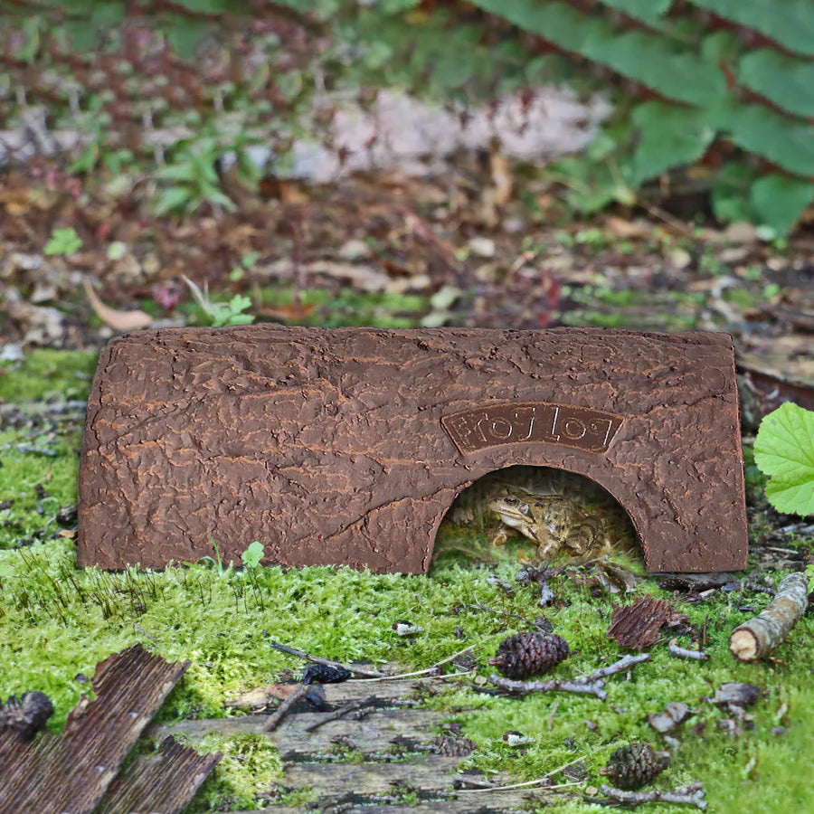 Simon King Frog Log, an eco-friendly amphibian shelter made from recyclable CLAYPLAS material, designed to resemble a realistic wooden log. A frog is seen resting inside the log, which is situated on mossy ground surrounded by natural woodland debris, offering a safe and shaded refuge for frogs and toads in wildlife-friendly gardens.