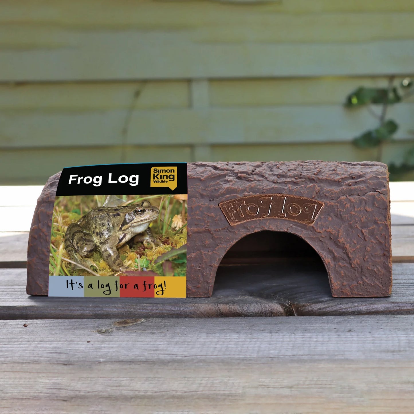 Simon King Frog Log, an eco-friendly amphibian shelter made from durable, recyclable CLAYPLAS material, resembling a realistic wooden log. Designed to protect frogs and toads from predators, the habitat features a natural appearance and is displayed on a wooden surface with product packaging showcasing a frog and the tagline 'It's a log for a frog!'