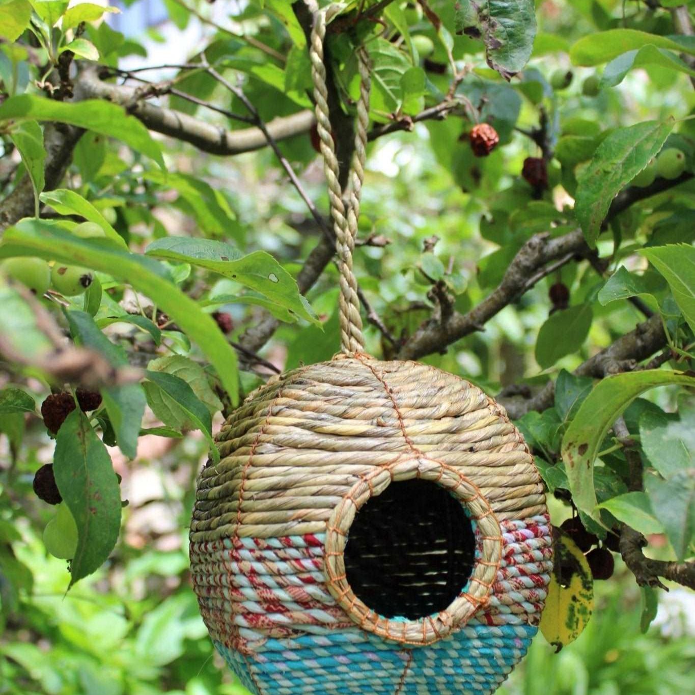 A spherical Anita Artisan Bird Nester woven from natural fibers, featuring colorful blue and red patterns, a round entrance hole, and a sturdy hanging rope, suspended from a tree branch surrounded by lush green foliage, designed as a cozy nesting space for small garden birds.






