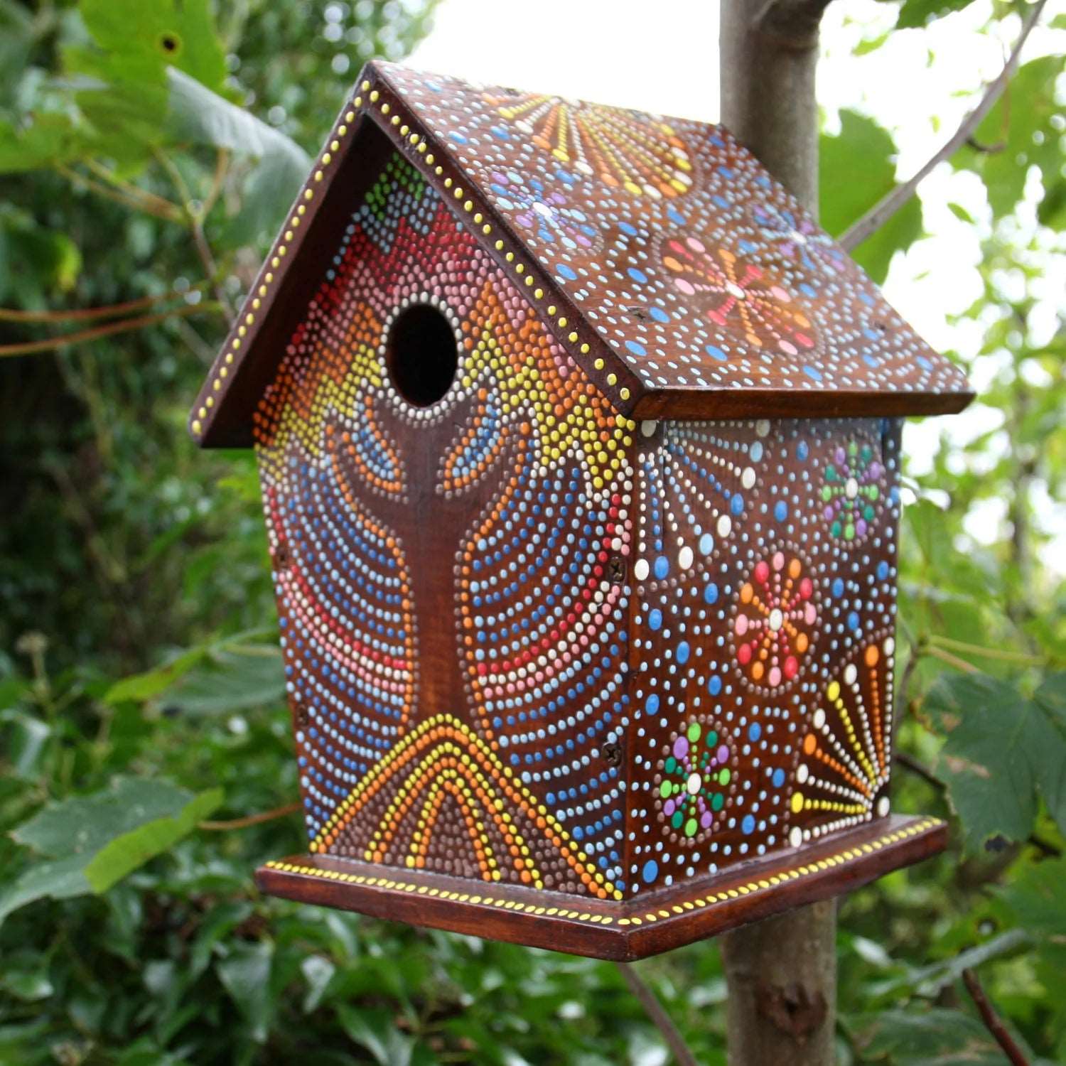 Colourful artisan birdhouse with intricate hand-painted patterns inspired by Bali Island designs, featuring vibrant dots and floral motifs set against a rich brown background, hanging on a tree branch surrounded by lush greenery.