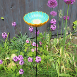 Ceramic African Daisy Bird Drinker, vibrant turquoise and yellow daisy-shaped bowl, mounted on a tall metal stand, surrounded by colorful purple and pink garden flowers, placed in a lush garden setting, ideal for attracting birds