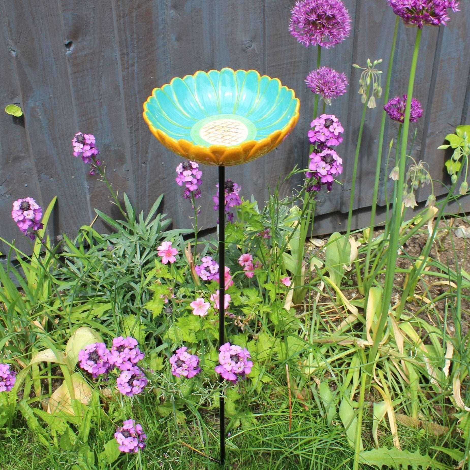 Ceramic African Daisy Bird Drinker, vibrant turquoise and yellow daisy-shaped bowl, mounted on a tall metal stand, surrounded by colorful purple and pink garden flowers, placed in a lush garden setting, ideal for attracting birds