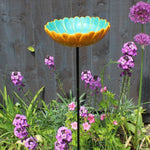 Ceramic African Daisy Bird Drinker, vibrant turquoise and yellow daisy-shaped bowl, mounted on a tall metal stand, surrounded by colorful purple and pink garden flowers, placed in a lush garden setting, ideal for attracting birds