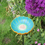 Ceramic African Daisy Bird Drinker, vibrant turquoise and yellow daisy-shaped bowl, mounted on a tall metal stand, surrounded by colorful purple and pink garden flowers, placed in a lush garden setting, ideal for attracting birds