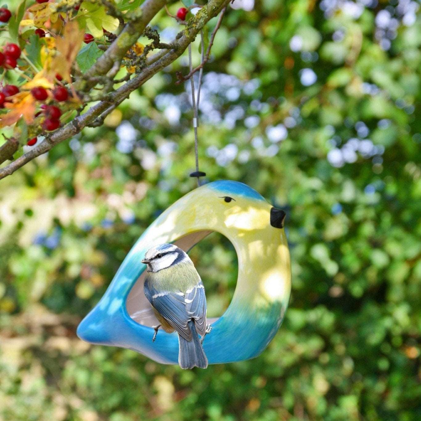 Ceramic Blue Tit Bird Feeder, vibrant blue and yellow bird-shaped feeder, blue tit perched at the entrance, surrounded by green foliage and red berries, hanging from a tree branch, eco-friendly garden accessory"