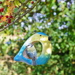 Ceramic Blue Tit Bird Feeder, vibrant blue and yellow bird-shaped feeder, blue tit perched at the entrance, surrounded by green foliage and red berries, hanging from a tree branch, eco-friendly garden accessory"