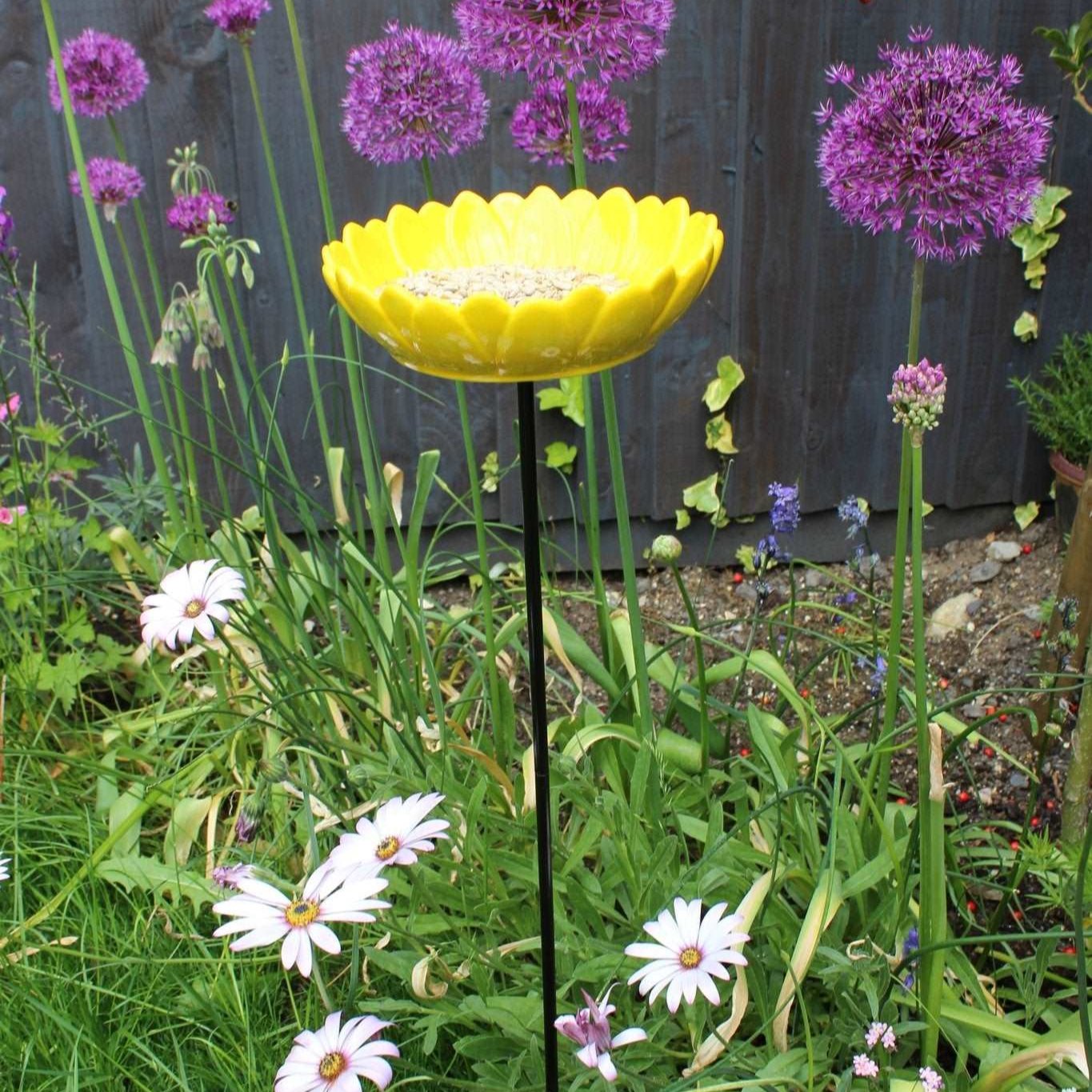 Bright yellow ceramic sunflower bird feeder mounted on a metal stake, filled with birdseed, placed in a vibrant garden setting with colorful flowers and greenery in the background.