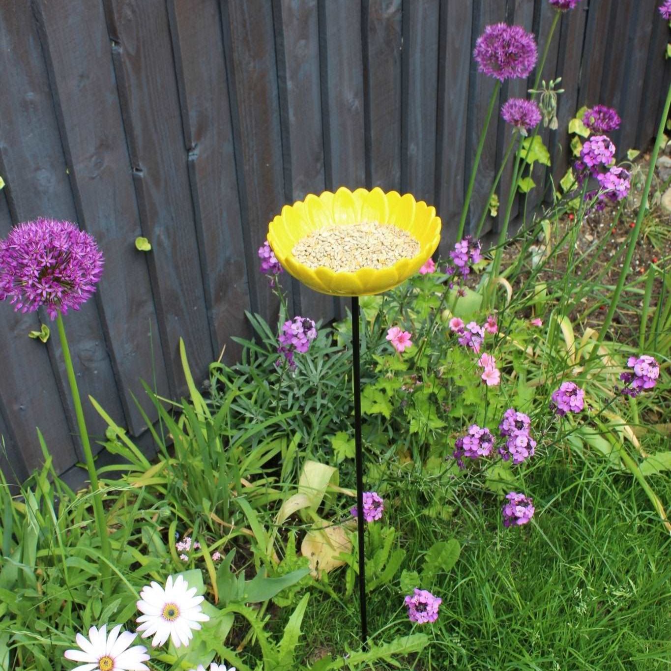 Bright yellow ceramic sunflower bird feeder mounted on a metal stake, filled with birdseed, placed in a vibrant garden setting with colorful flowers and greenery in the background.