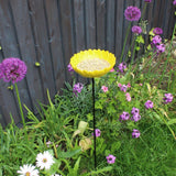 Bright yellow ceramic sunflower bird feeder mounted on a metal stake, filled with birdseed, placed in a vibrant garden setting with colorful flowers and greenery in the background.