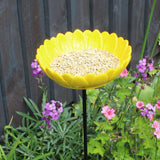 Bright yellow ceramic sunflower bird feeder mounted on a metal stake, filled with birdseed, placed in a vibrant garden setting with colorful flowers and greenery in the background.