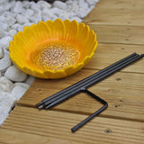 Disassembled ceramic sunflower bird feeder showing the vibrant yellow sunflower-shaped bowl placed on a wooden deck next to the black metal stake, with white decorative stones in the background