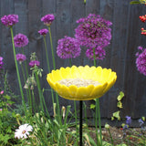 Bright yellow ceramic sunflower bird feeder mounted on a metal stake, filled with birdseed, placed in a vibrant garden setting with colorful flowers and greenery in the background.