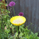 Bright yellow ceramic sunflower bird feeder mounted on a metal stake, filled with birdseed, placed in a vibrant garden setting with colorful flowers and greenery in the background.