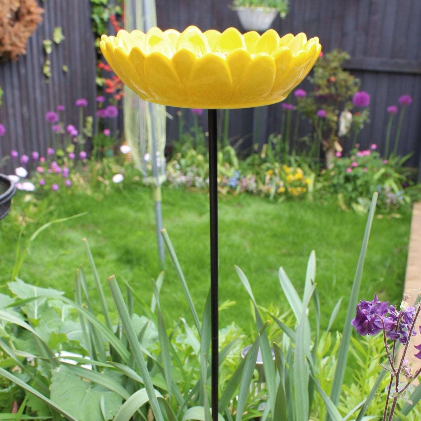 Bright yellow ceramic sunflower bird feeder mounted on a metal stake, filled with birdseed, placed in a vibrant garden setting with colorful flowers and greenery in the background.