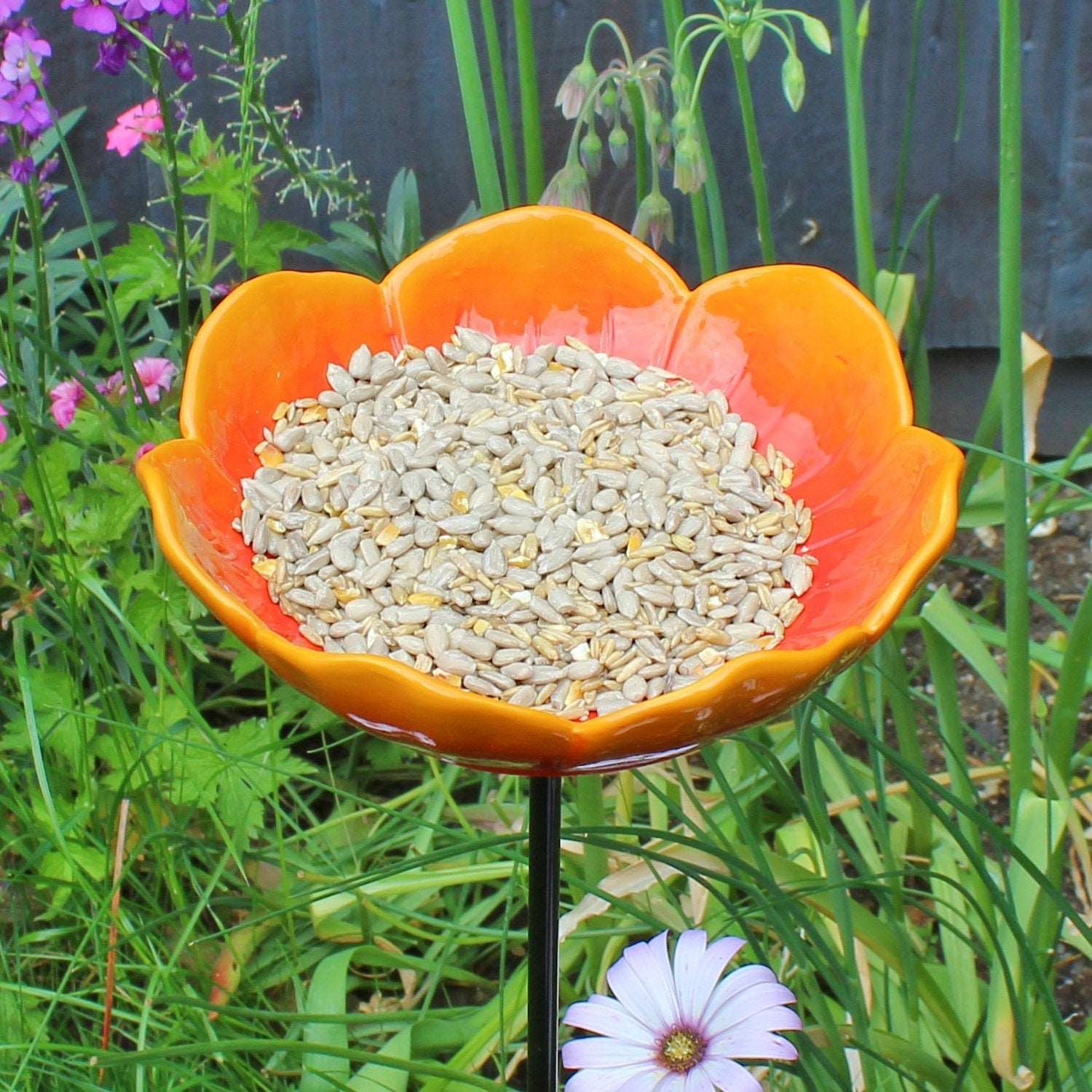 Bright orange ceramic wild poppy-shaped bird feeder filled with sunflower seeds, placed on a black metal pole, surrounded by vibrant green grass and colorful garden flowers