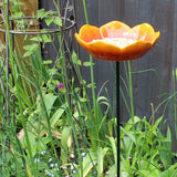 Bright orange ceramic wild poppy-shaped bird feeder filled with sunflower seeds, placed on a black metal pole, surrounded by vibrant green grass and colorful garden flowers