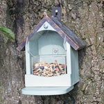 Craftsman squirrel feeder mounted on a tree trunk, featuring a house-shaped design with a slanted metal roof, , clear acrylic seed compartment filled with nuts and seeds, and a front perch for easy access