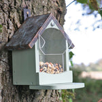 Craftsman squirrel feeder mounted on a tree trunk, featuring a house-shaped design with a slanted metal roof, light green wooden frame, clear acrylic seed compartment filled with nuts and seeds, and a front perch for easy access