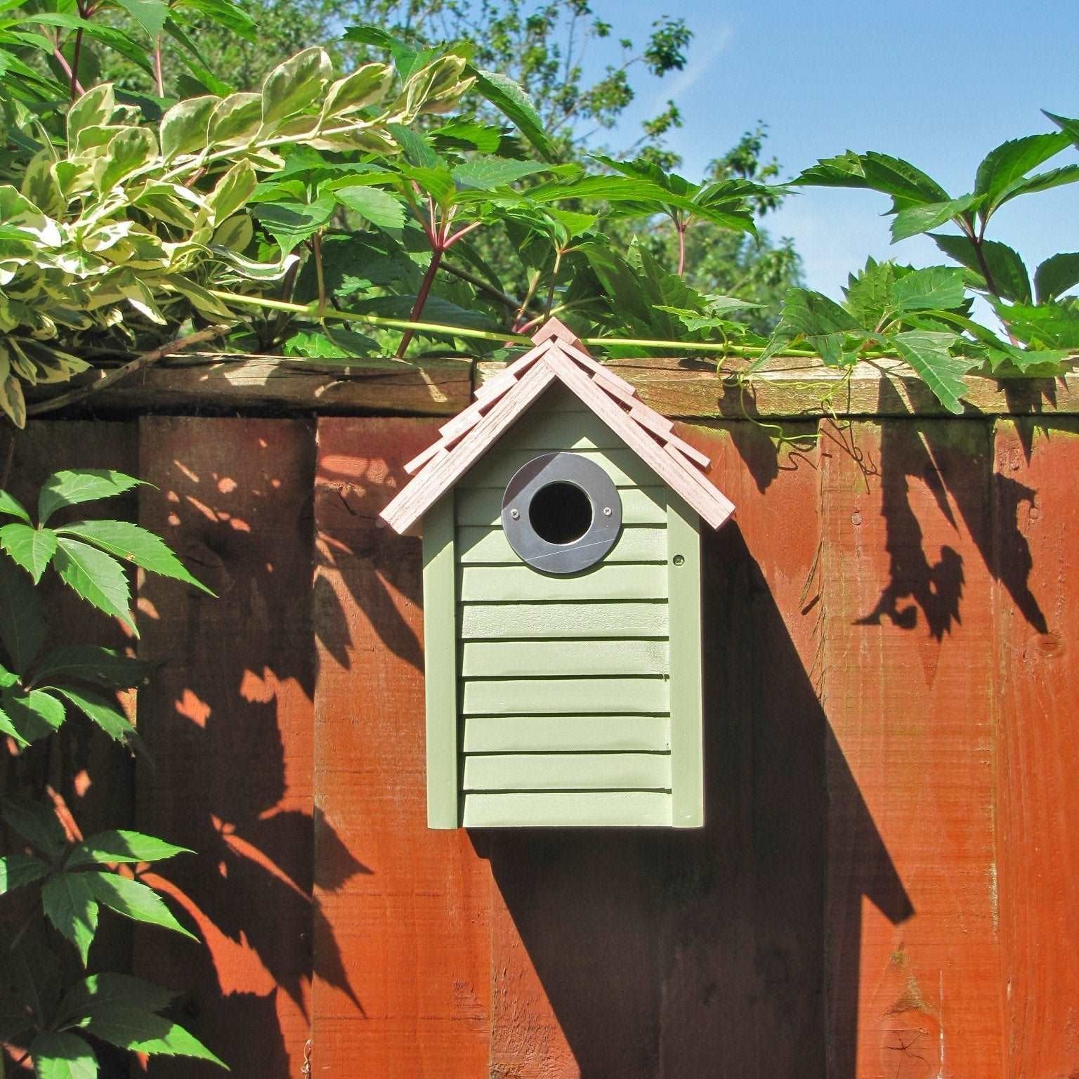 New England Nest Box - The Irish Gardener Store New England Nest Box, green wooden birdhouse with horizontal slats, natural wood-colored pitched roof, circular black metal entrance hole, mounted on a wooden fence, surrounded by lush green foliage, durable and stylish bird nesting box for garden birds