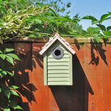 New England Nest Box - The Irish Gardener Store New England Nest Box, green wooden birdhouse with horizontal slats, natural wood-colored pitched roof, circular black metal entrance hole, mounted on a wooden fence, surrounded by lush green foliage, durable and stylish bird nesting box for garden birds